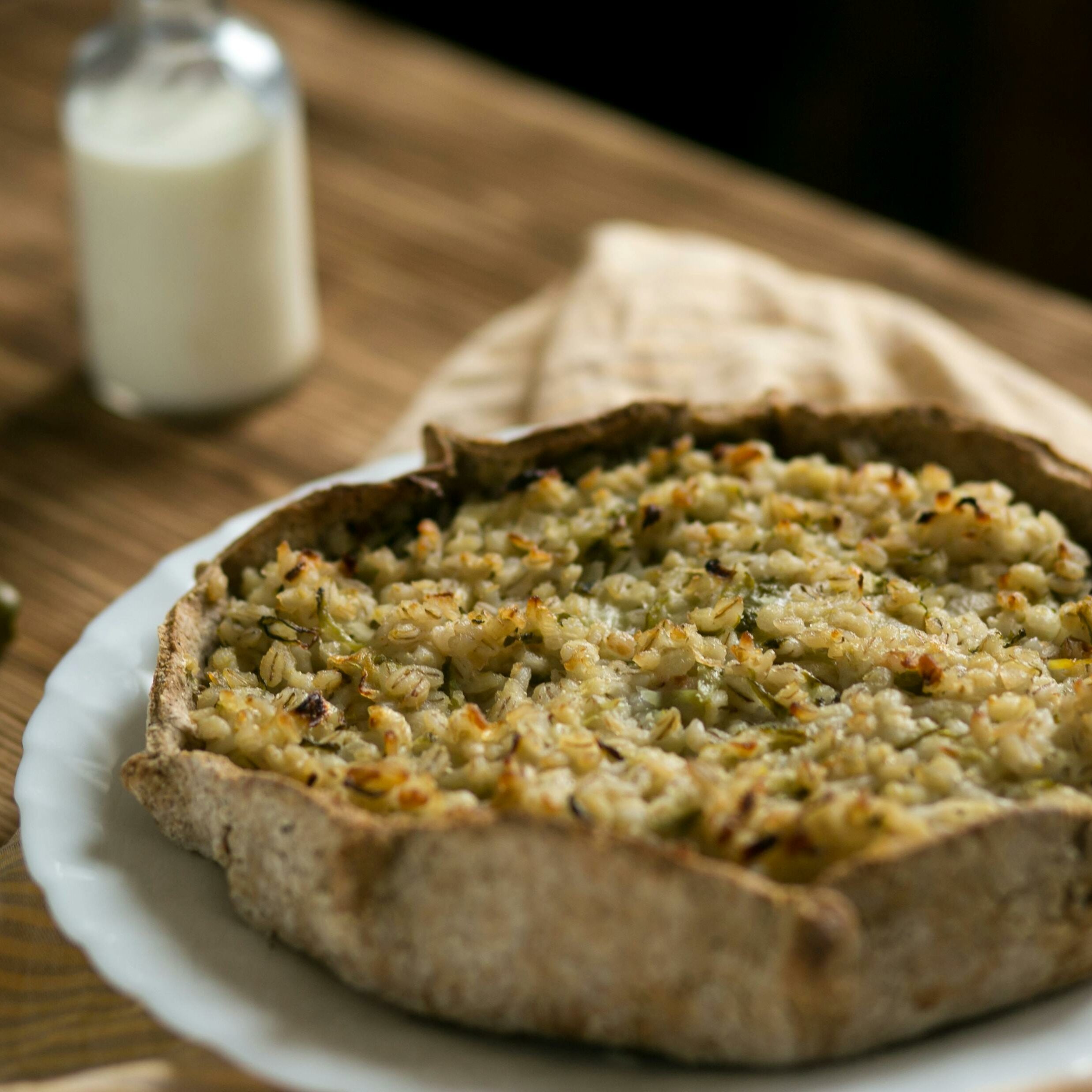 Tarte rustique aux céréales et légumes, cuite au four avec une pâte complète, servie sur une assiette blanche en bois, accompagnée d'une bouteille de lait végétal en arrière-plan.