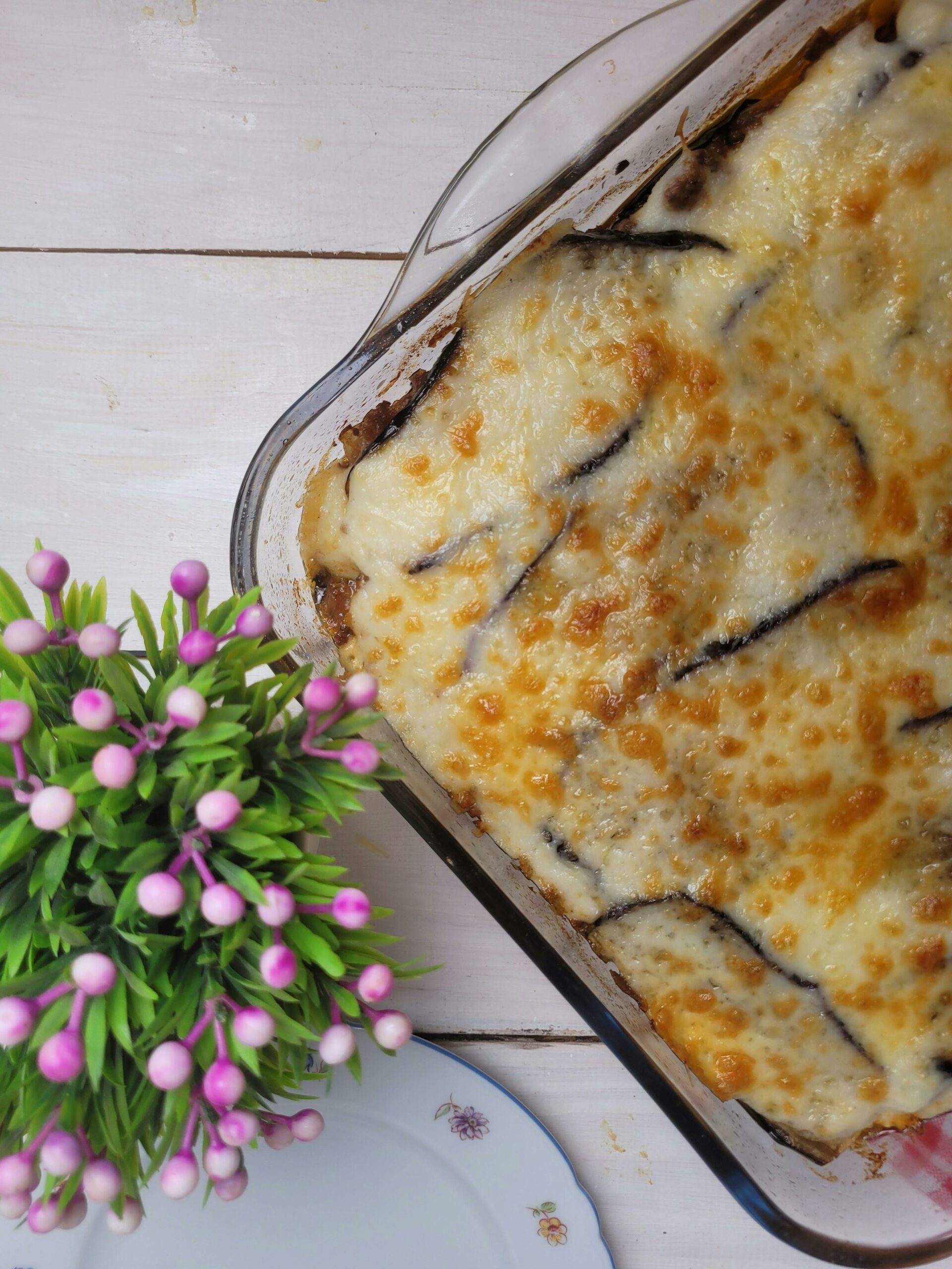 Gratin d’aubergines au fromage gratiné dans un plat en verre, avec une croûte dorée et fondante, posé sur une table en bois blanc à côté d’une assiette fleurie et d’une plante décorative aux fleurs roses.